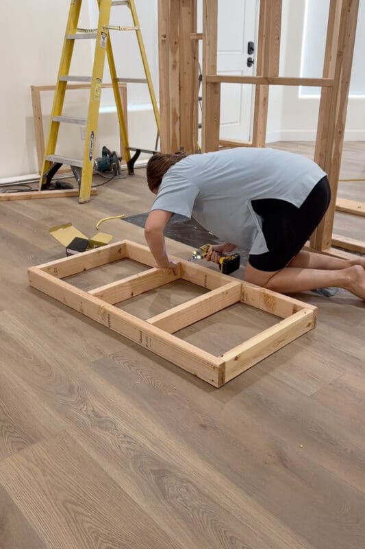 Crystel kneeling on the floor using an impact driver to assemble a small 2x4 frame that will fill the space above the arched doorway