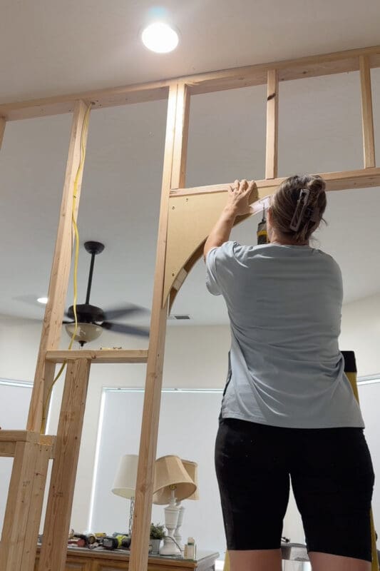 Crystel on a ladder using a drill to secure the bottom of an MDF arch piece to the existing bedroom wall next to a wood barn door