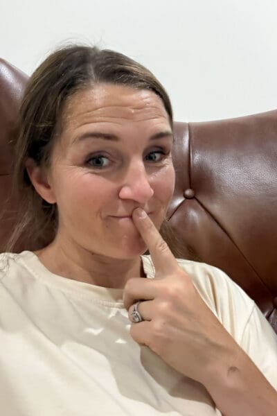 Crystel smiling with a finger to her lips and a playful expression while sitting in a brown leather chair