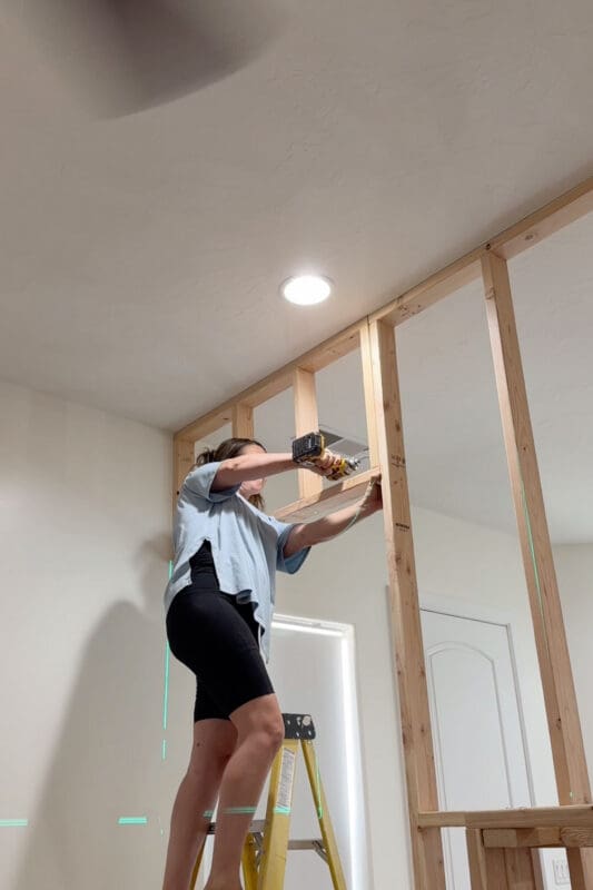 Crystel standing on a ladder screwing a 2x4 frame into place below the ceiling to lower the top of the wall opening for the arch