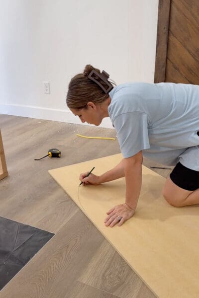 Crystel kneeling on the floor freehand sketching a curved arch line in pencil on a large sheet of board, with a tape measure nearby