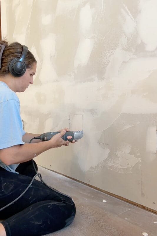 Crystel wearing ear protection and using a power tool to cut a small rectangular opening in the drywall on the back side of the fireplace wall