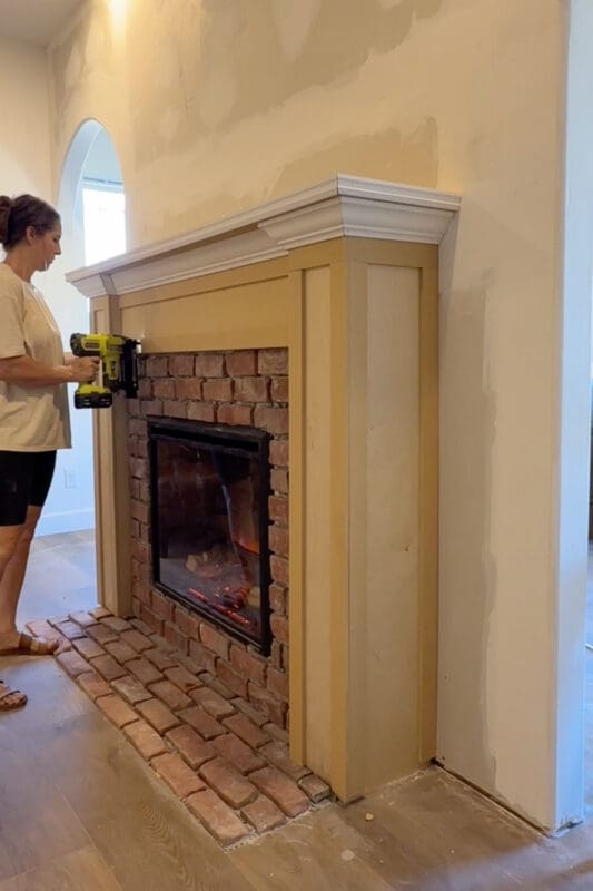 Crystel using a cordless brad nailer to attach MDF trim to the side column of the fireplace surround, beside the brick and glowing electric insert