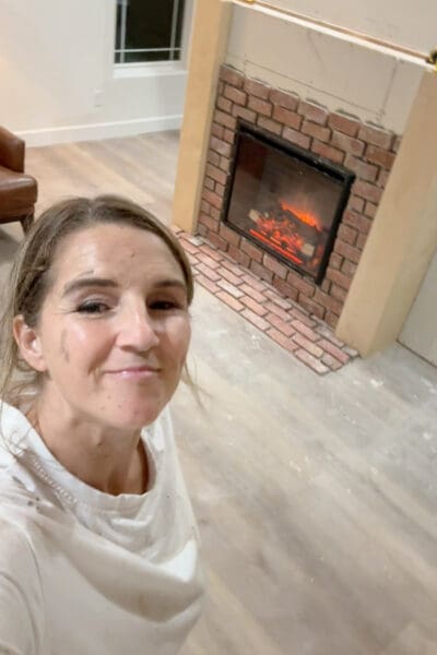 Crystel taking a smiling, mortar-smudged selfie in front of the electric fireplace after finishing the brick surround and hearth, with the insert glowing and the plywood side columns in place