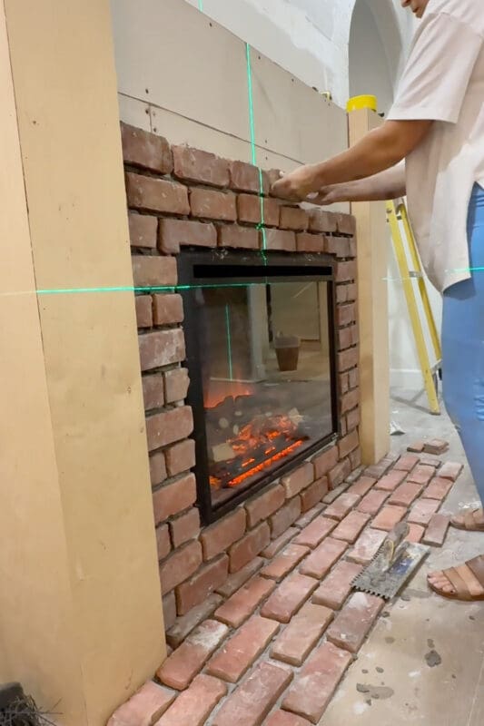 Crystel setting red bricks around the electric fireplace insert using a green laser level as a guide, with the finished brick hearth and a notched trowel on the floor below