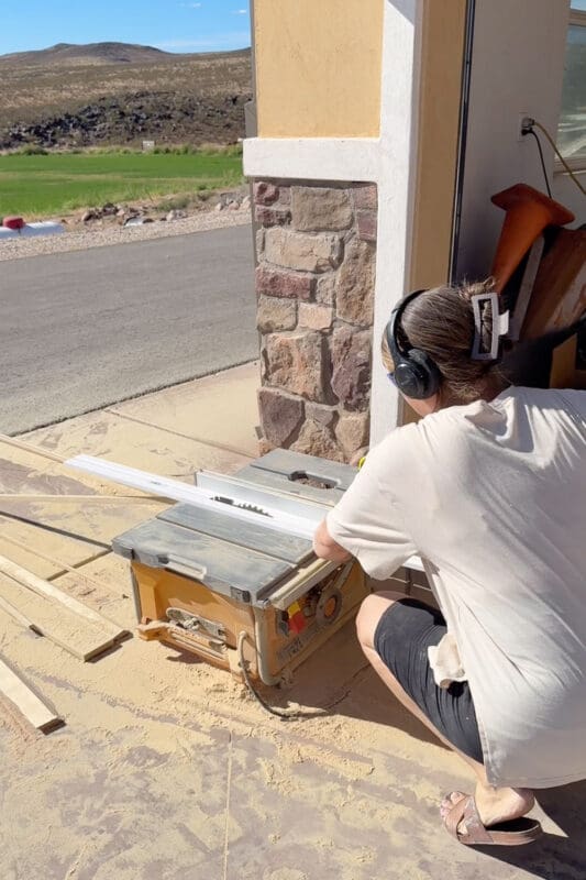 Crystel wearing ear protection and using a table saw outside to rip a piece of baseboard down into trim for the mantel edge