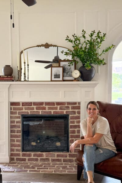 Finished electric fireplace built from scratch with a red brick surround and hearth, a white mantel decorated with brass candlesticks, framed photos, and a vase of greenery, and Crystel seated beside it near an arched doorway