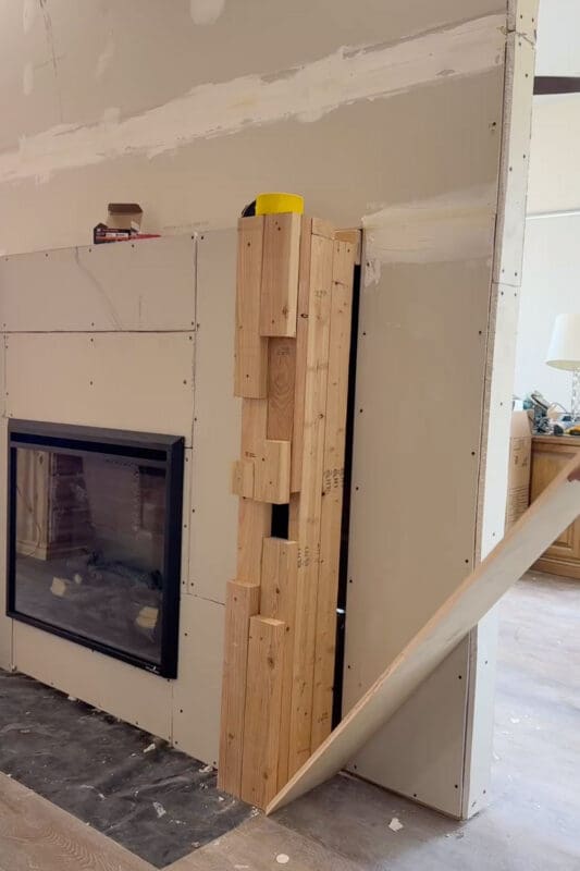 Electric fireplace insert installed in the drywalled recess, with stacked wood blocks framing out the side column and a sheet of plywood being fitted against it