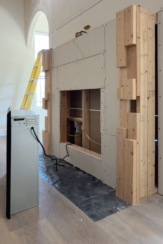 Electric fireplace insert standing beside the drywalled recess with its power cord running to the outlet inside the opening, ready to be slid into place