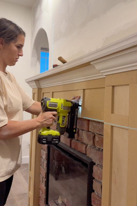 Crystel nailing MDF trim pieces onto the mantel front above the brick to create box molding panels, using a laser level to keep the lines even