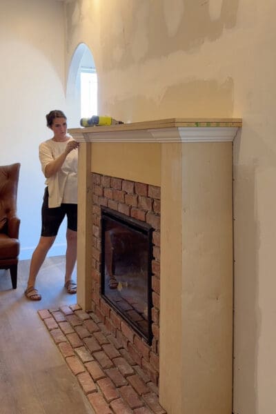Crystel checking the crown molding installed beneath the mantel shelf on the plywood fireplace surround, with the brick hearth in front