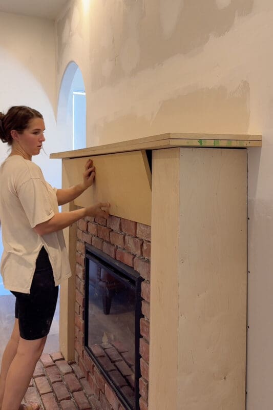 Crystel fitting a plywood panel onto the front of the mantel above the brick, set slightly back from the two side columns, beneath the double-stacked plywood shelf