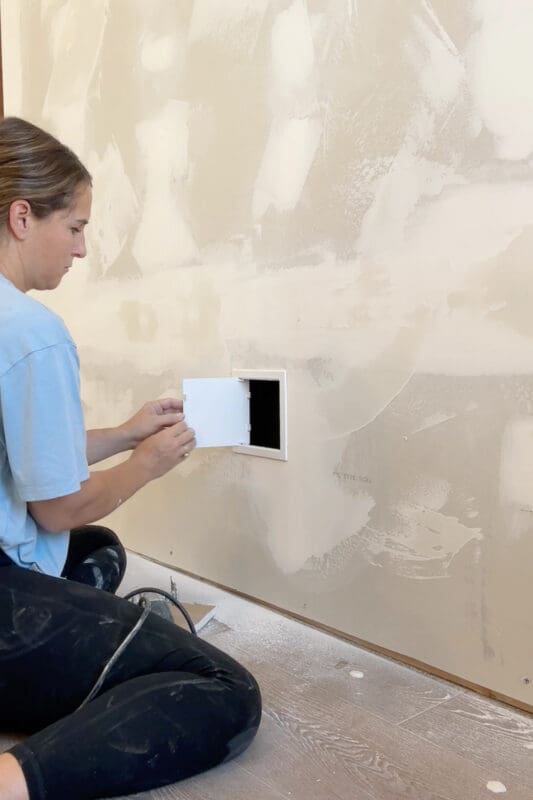 Crystel attaching a small white access door to the drywall cutout on the back of the wall, giving easy access to the electric fireplace outlet