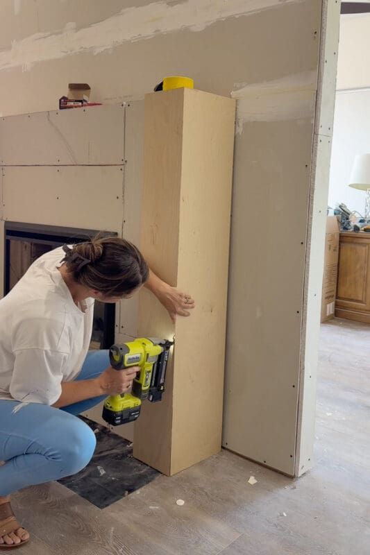 Crystel using a cordless brad nailer to attach a plywood panel that wraps the side column of the fireplace surround