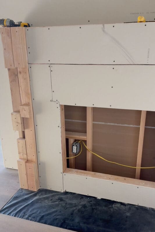 Close-up of the drywalled fireplace surround with an outlet and yellow wire inside the recessed opening, ready for the electric insert, and plastic sheeting on the floor where the hearth will go