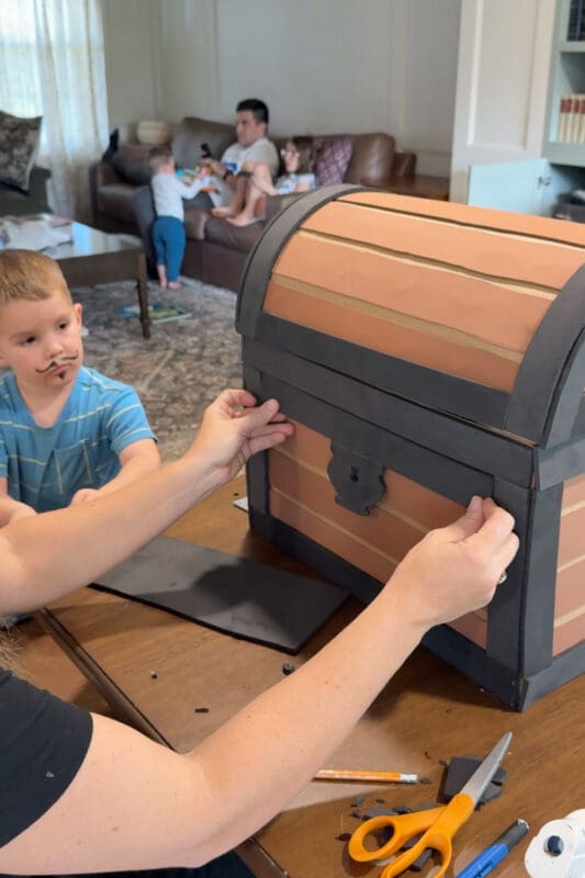 Crystel pressing a black craft foam band into place along the front of the cardboard treasure chest, with the foam lock plate centered just below the lid, as a young helper watches from beside the table.