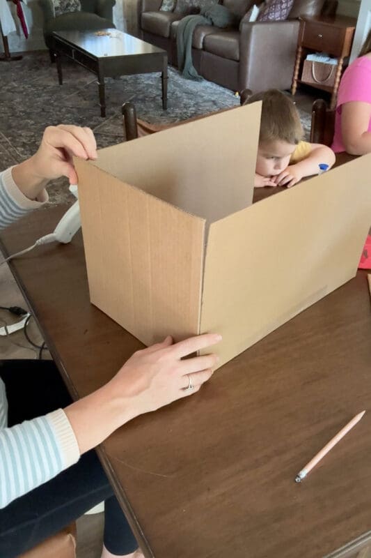 Crystel holding a cardboard side panel in place against the front and back pieces of a custom-sized cardboard box base, lining up the edges before gluing, while a young helper rests their chin on the table behind it.