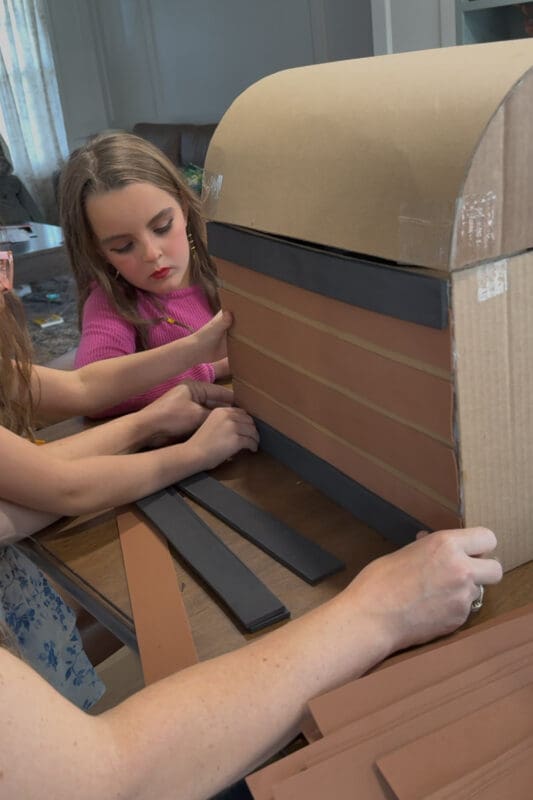 Crystel pressing a thinner strip of black craft foam along the bottom edge of the DIY treasure chest to mimic metal trim, with a matching black strip already framing the top edge and a young helper watching closely.