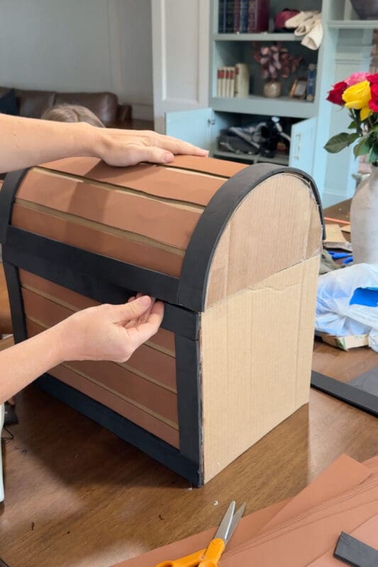 Crystel pressing black craft foam trim along the front edge of a DIY treasure chest now covered in brown foam planks on both the lid and base, with the rounded cardboard side still bare.