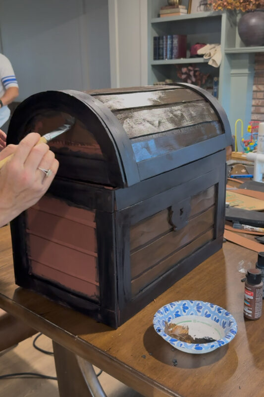 Crystel painting brown acrylic over the planks on the rounded side of the cardboard treasure chest lid, with the unpainted reddish primer still showing on the side of the base below for a clear before and after.