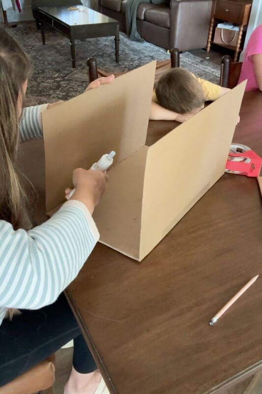 Crystel holding the front and back cardboard panels upright while hot gluing the second inside seam to form the open base of a pirate treasure chest, with a young helper resting their head on the table behind it.