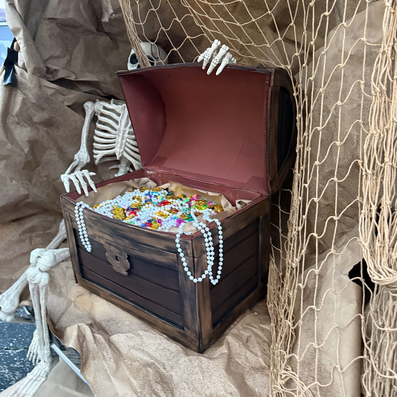 Close-up of a cardboard treasure chest painted to look like aged wood, its curved lid open to a reddish-brown interior, with pearl necklaces draped over the front, a skeleton hand gripping the lid, and fishing net hanging alongside.