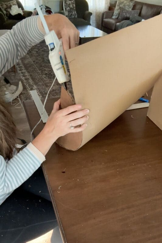 Crystel bending the flat cardboard over the rounded side piece and hot gluing it in place as she goes, shaping the curved top of a cardboard chest lid.