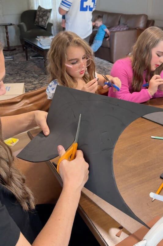 Crystel cutting a curved arch out of black craft foam with orange-handled scissors, following lines traced from the same mixing bowl used for the lid sides, while young helpers trim their own foam scraps at the table.