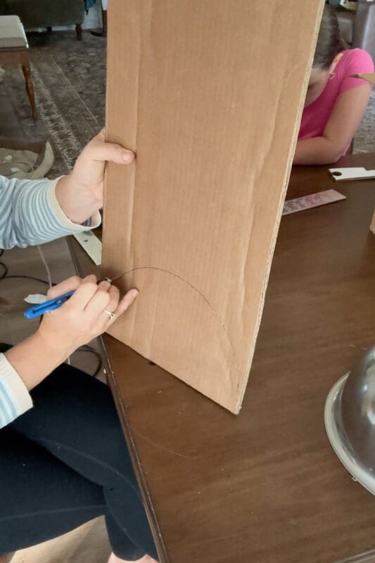 Crystel holding the cardboard upright and cutting along the traced curved line with a blue utility knife to make a rounded side piece for a cardboard chest lid, as a young helper works at the table behind her.