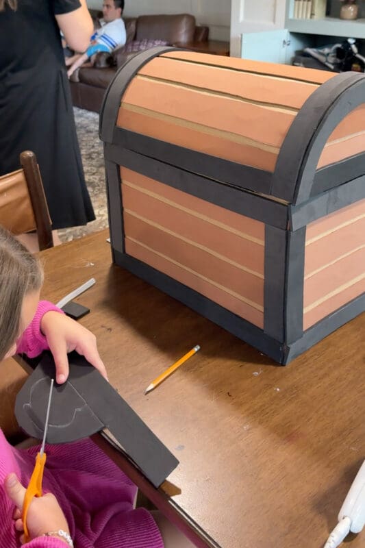 A young helper cutting a traced lock plate shape out of black craft foam with orange-handled scissors, next to the fully planked and trimmed DIY treasure chest.