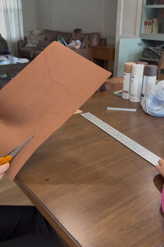 Cutting brown craft foam along penciled lines with orange-handled scissors to make faux wood planks for a cardboard treasure chest, while a young helper holds a metal ruler on the table.