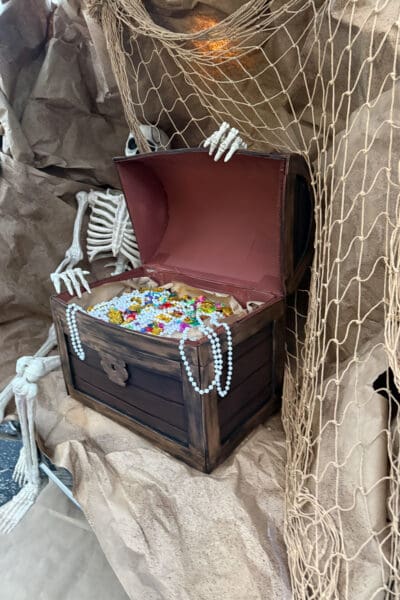 Finished DIY treasure chest with an aged gold trimmed lid propped open, overflowing with gold coins, colorful gems, and pearl strands, while a skeleton reaches over the edge and fishing net hangs beside it.