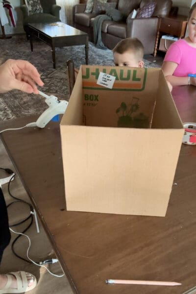 Finished open cardboard box sitting on a wood table, with printing from a repurposed moving box visible on the inside panel and two young helpers peeking over the top.