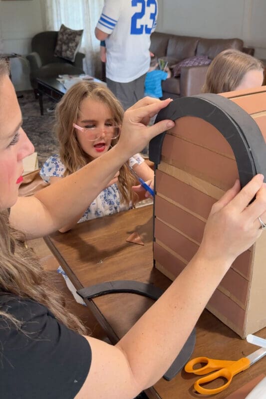 Crystel fitting a rounded black craft foam arch over the planked side of the cardboard treasure chest lid to frame the curve like metal banding, as a young helper looks on from across the table.