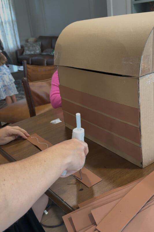 Crystel running a line of hot glue down a brown craft foam strip before pressing it onto the front of a cardboard treasure chest, where three foam planks are already lined up with small gaps between them to look like wood boards.