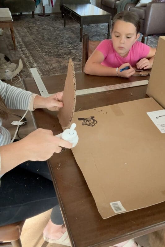 Crystel hot gluing the curved edge of a rounded cardboard side piece onto a flat sheet of cardboard cut to the width of the chest, as a young helper watches from across the table.