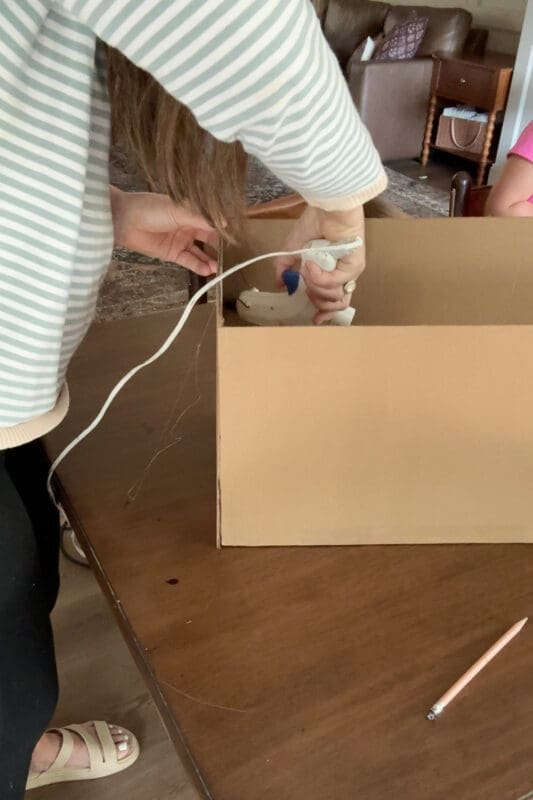 Crystel reaching into the cardboard box with a hot glue gun to seal the inside seam where the side panel meets the front of her cardboard treasure chest.