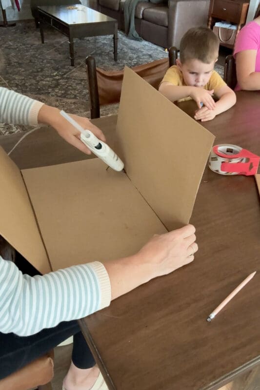 Crystel folding up one side of the taped cardboard and running hot glue along the inside seam to start shaping a cardboard box, as a young helper watches from across the table.