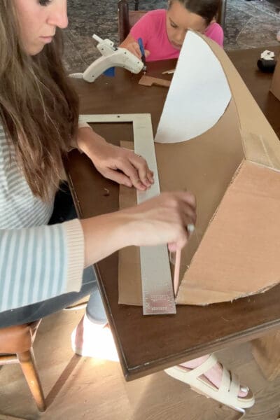 Crystel lining up a metal ruler and marking with a pencil where to trim the extra cardboard length off the curved pirate treasure chest lid, while a young helper works at the table behind her.