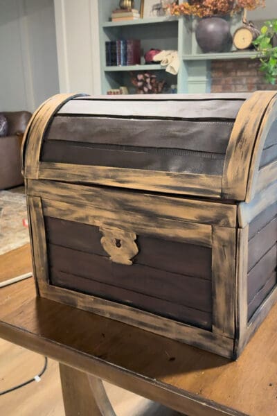 Front of the cardboard treasure chest after the Rub ‘n Buff step, with streaky aged gold metallic trim and a matching lock plate framing the dark brown painted foam planks.