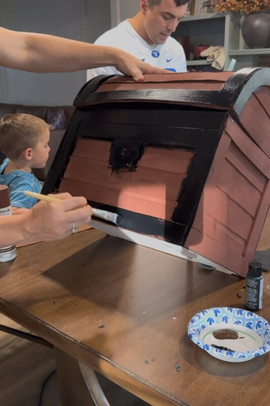 Crystel tipping the primed DIY treasure chest back and brushing black acrylic craft paint onto the raised foam trim along the bottom edge, with the front banding and lock already painted black and a young helper sitting nearby.