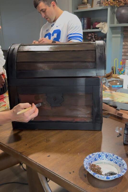 Crystel brushing dark brown acrylic paint over the front foam planks of the DIY treasure chest, leaving streaky brushstrokes that read like wood grain, with the black trim and lock already finished.