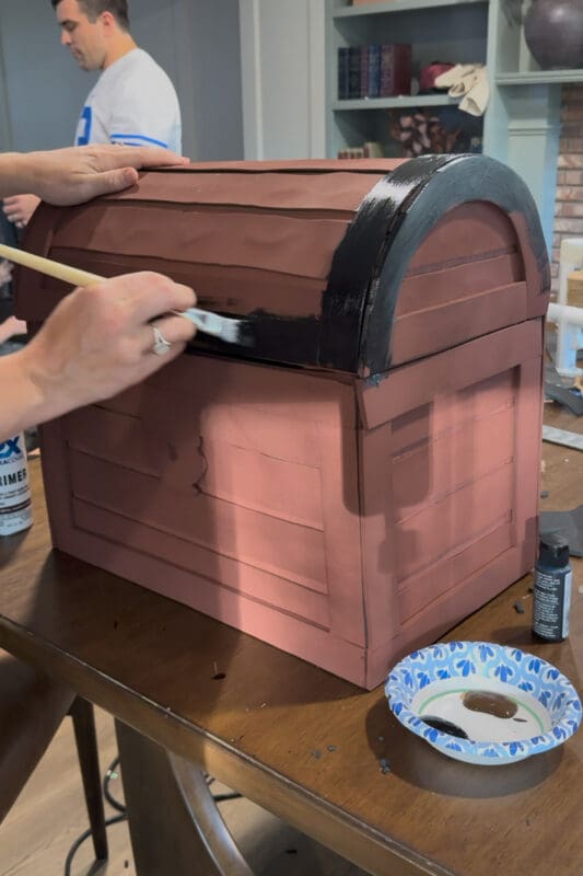 Crystel brushing black acrylic paint along the raised foam trim on the front of the curved lid, with the rounded side banding already painted and a paper bowl of black and brown paint on the table beside the cardboard treasure chest.