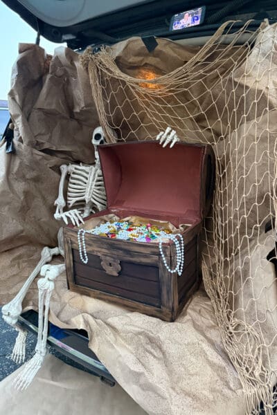 DIY treasure chest with a weathered dark wood finish sitting open in a van’s cargo area, filled with gold coins, colorful gems, and pearl strands, while a skeleton peeks over the lid and a phone streams a football game from the liftgate above a pirate trunk or treat setup.