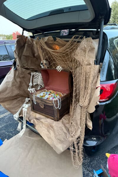 Full pirate trunk or treat setup in an open van, with crumpled brown paper lining the cargo area, a draped fishing net, a skeleton sprawled beside the open cardboard treasure chest, and paper spilling down over the bumper onto the parking lot.