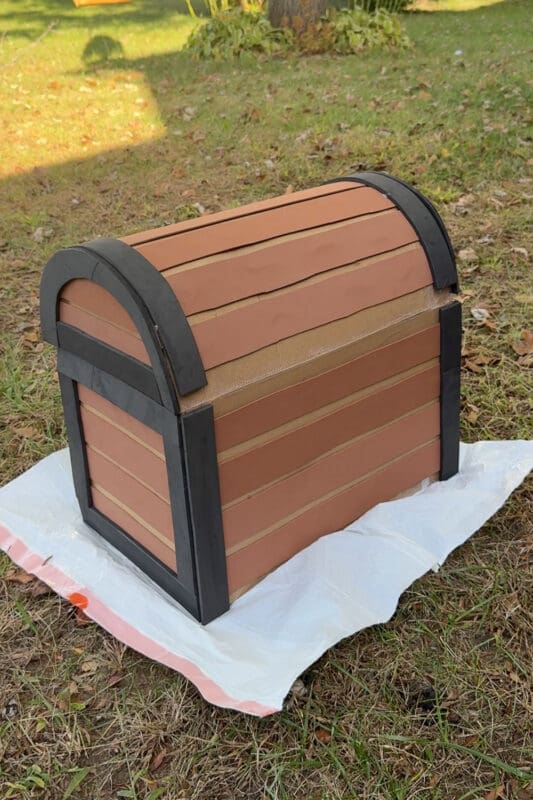 Back view of the finished cardboard and craft foam DIY treasure chest sitting on a plastic drop cloth in the yard before painting, with a strip of bare cardboard left uncovered along the hinge so the lid can open freely.