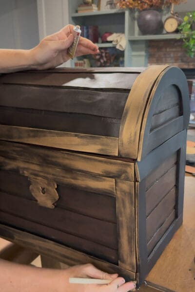 Crystel holding a tube of Rub ‘n Buff and rubbing the metallic wax onto the black foam trim with her fingers, giving the front banding and lock of the DIY treasure chest a worn gold finish while the side trim is still plain black.