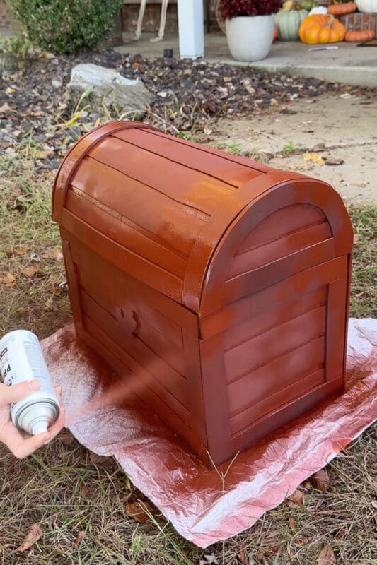 Crystel spraying red primer over the entire cardboard treasure chest outdoors on a drop cloth, covering the foam planks and trim in one even reddish-brown coat, with fall pumpkins on the porch behind it.