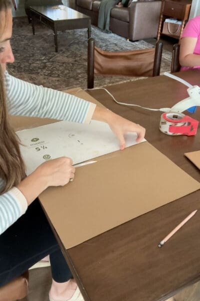 Crystel running clear packing tape along the seam between equal cardboard rectangles laid flat on a wood table, temporarily joining the bottom, front, and back pieces of a custom sized cardboard box.