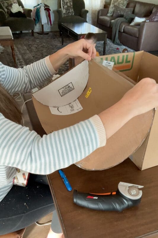 Crystel holding the cardboard treasure chest lid open and smoothing packing tape along the inside of the hinge seam to reinforce it, as a young helper peeks over the box from behind.
